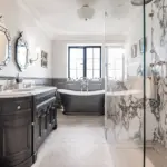 A bathroom featuring a black double vanity with a marble worktop, a freestanding black bath centred under a window, and a glass-enclosed shower with marbled tiled walls.