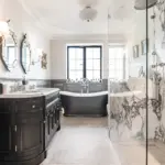 A luxury bathroom featuring a dark vanity with a marble worktop, a freestanding black bath centred under a window, and a glass-enclosed shower with marble walls.