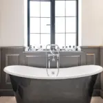 A freestanding black clawfoot bath with silver fittings sits before a window lined with glass perfume bottles.