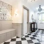 A bright entrance hall featuring a built-in bench, panelled wainscotting, a diamond-patterned mirrored wall, and a black-and-white chequerboard tiled floor.