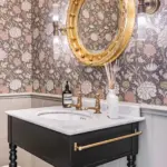 A bathroom vanity with a marble top, gold tap, and circular gold mirror, set against floral wallpaper.