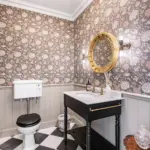 A bathroom featuring floral wallpaper, a toilet with a dark seat, a black vanity with a marble top and gold fittings, a circular gold mirror, and black and white chequerboard flooring.
