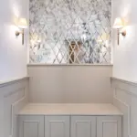 A built-in grey bench with storage cupboards sits beneath a large, diamond-patterned antique mirror, flanked by two wall lights above white panelling.