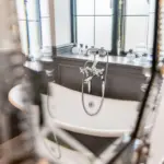 A white clawfoot bath with chrome fittings placed in front of a window, seen through a blurred ornate mirror frame.