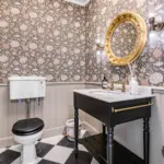 A small bathroom with floral wallpaper, black and white chequered floors, a toilet with a wall-mounted cistern, and a black vanity with a gold-framed circular mirror.