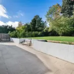 A paved patio with a single wooden chair next to a low white retaining wall and a raised lawn.