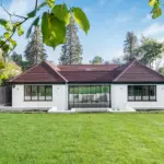 A white single-storey house with a dark tiled roof and a large central glass door, seen across a green lawn.