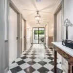 A brightly lit corridor with a black-and-white chequered marble floor leading towards a glass-panelled door, with a console table and decorative items in the foreground.