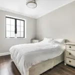 A bedroom with a bed featuring a white grid-patterned duvet, flanked by two antique-style white chests of drawers near a window.