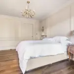 A bedroom with pale walls, a white-covered bed, a chandelier, and a wooden bedside table next to an upholstered chair.