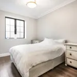 A bedroom with a bed covered in a white quilt, a window, and a wooden chest of drawers.