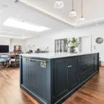 A dark blue kitchen island with a white worktop and brass fittings sits in an open-plan kitchen and living area with wooden flooring.