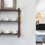 A wooden wall shelf holding six floral teacups on saucers, with a framed mirror above and a glimpse into a sitting room in the background.