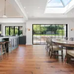 A spacious kitchen and dining area with a dark grey kitchen island, three wooden stools, a long wooden dining table, and large black-framed glass doors leading to a patio.
