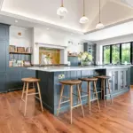 A spacious kitchen with dark blue cabinetry, a large white-topped island with three wooden stools, and wooden flooring.
