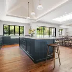 A spacious kitchen with a dark blue central island and cabinetry, wooden flooring, and a dining area with a long table and chairs.