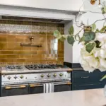 A large, professional-style stainless steel gas cooker with gold knobs sits beneath a brass pot filler tap mounted on a wall of glossy, mustard-coloured metro tiles.