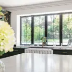 A kitchen sink with a gold tap is positioned in front of a window with black frames, overlooking a back garden.