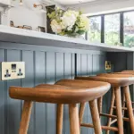 Three wooden bar stools positioned against a dark green kitchen island with brass electrical sockets.