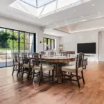 A dining area with a large circular wooden table surrounded by eight chairs, situated under a skylight next to a wall of floor-to-ceiling windows looking out onto a patio.