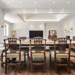 A dining room with a long oval wooden table surrounded by eight wooden chairs with spindle backs, facing a television mounted on a wall above a fireplace.