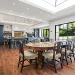 A dining room with a large oval wooden table and six chairs, adjacent to a kitchen with dark cabinetry, a central island, and large glass sliding doors opening to a patio.