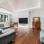 A living room with a large grey corner sofa, a wall-mounted television, a wooden drinks cabinet, and a fireplace with a patterned tiled hearth.