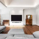 A spacious living room featuring a wall-mounted television, a grey corner sofa, a fireplace, and a tall gold-framed shelving unit.