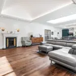 A spacious living room with dark hardwood floors, a grey leather corner sofa, a fireplace with decorative tiles, and an open-plan kitchen in the background.