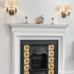 White fireplace mantel featuring a black cast-iron insert with floral tiles, two brass wall sconces, and a figurine of three cherubs.