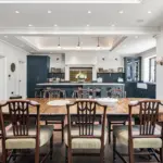 A dining room with a long wooden table surrounded by eight chairs, opening into a kitchen with dark blue cupboards, a central island, and a tiled splashback.