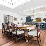 A large wooden dining table with upholstered chairs sits in a bright kitchen with dark cupboards, a centre island, and a skylight.