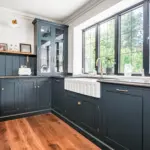 A kitchen with dark teal cupboards, a white butler sink, a brass tap, and a large window looking out onto a garden.