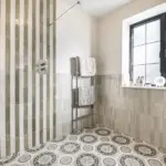 A walk-in shower with vertical striped tiled walls, patterned floor tiles, and a chrome towel radiator placed beneath a window.