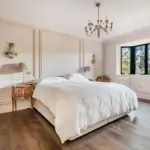 A bedroom with a central bed featuring a white duvet, flanked by two wooden bedside tables with lamps, against a wall with panelled moulding and a bay window.