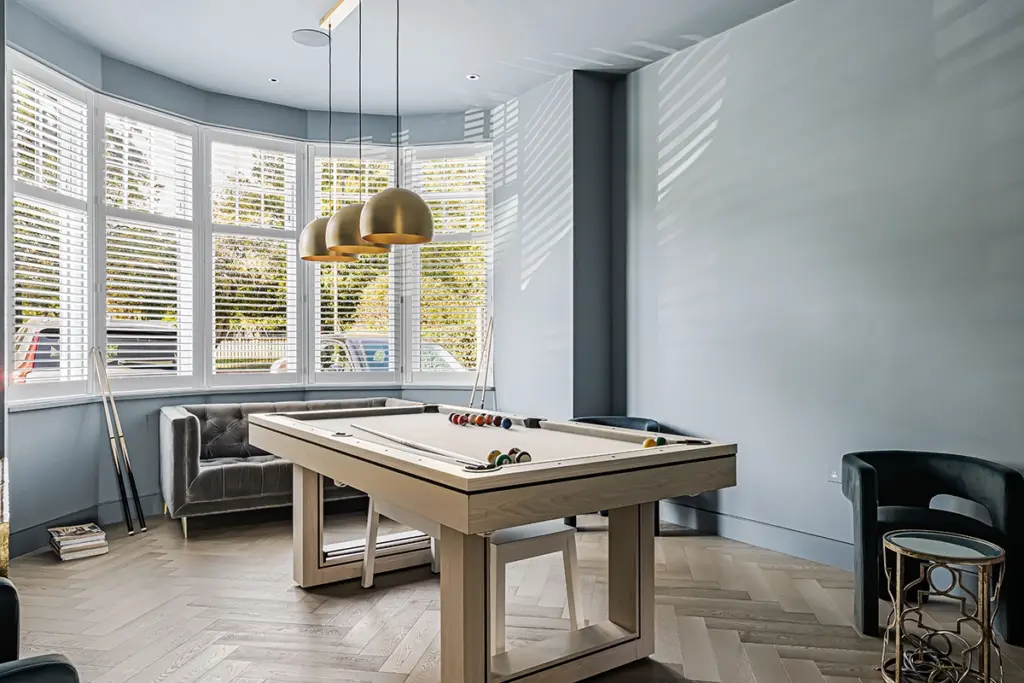 A light wood pool table sits in the centre of a blue-walled room with bay windows, hanging brass pendant lights, and a grey velvet sofa.