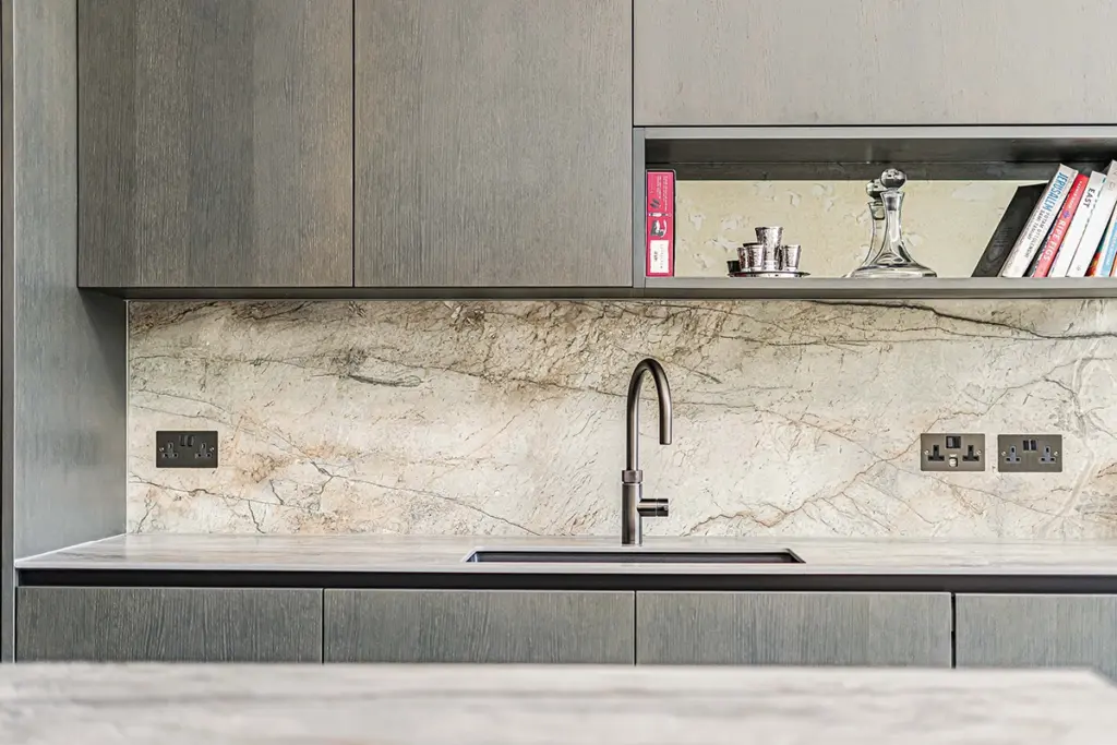A kitchen sink with a dark tap set against a marble splashback, with dark wood cupboards above and below and an open shelf containing books and glassware.