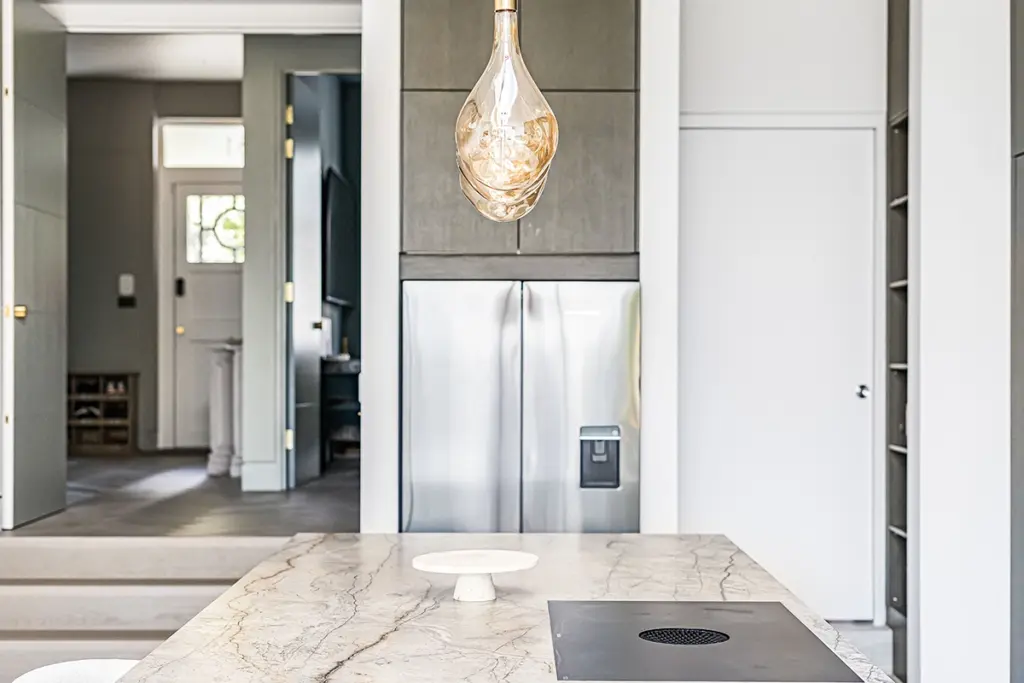 A kitchen island with a marble worktop, an induction hob, and a small white cake stand, positioned beneath a pendant light with a stainless steel fridge in the background.
