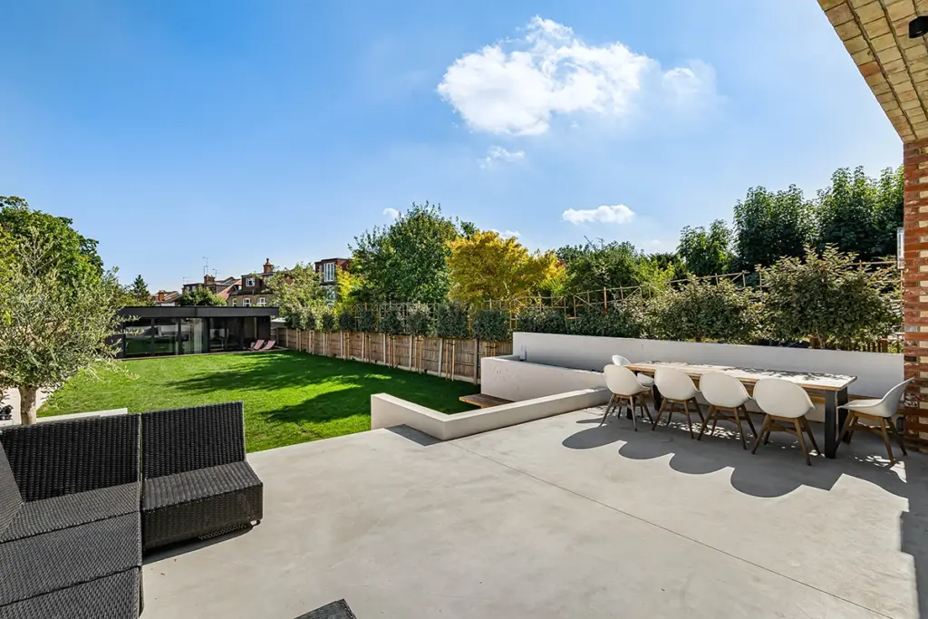 A patio featuring a large dining table with white chairs and black wicker lounge seating, overlooking a lawn with a garden room in the distance.