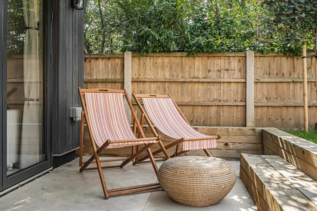 Two wooden deckchairs with striped fabric cushions and a woven round pouffe on a patio.
