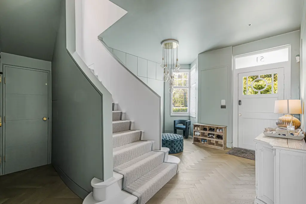 An entrance hall with light green walls, a staircase with a grey patterned runner, a white front door, a shoe rack, a blue velvet ottoman, and a blue armchair.