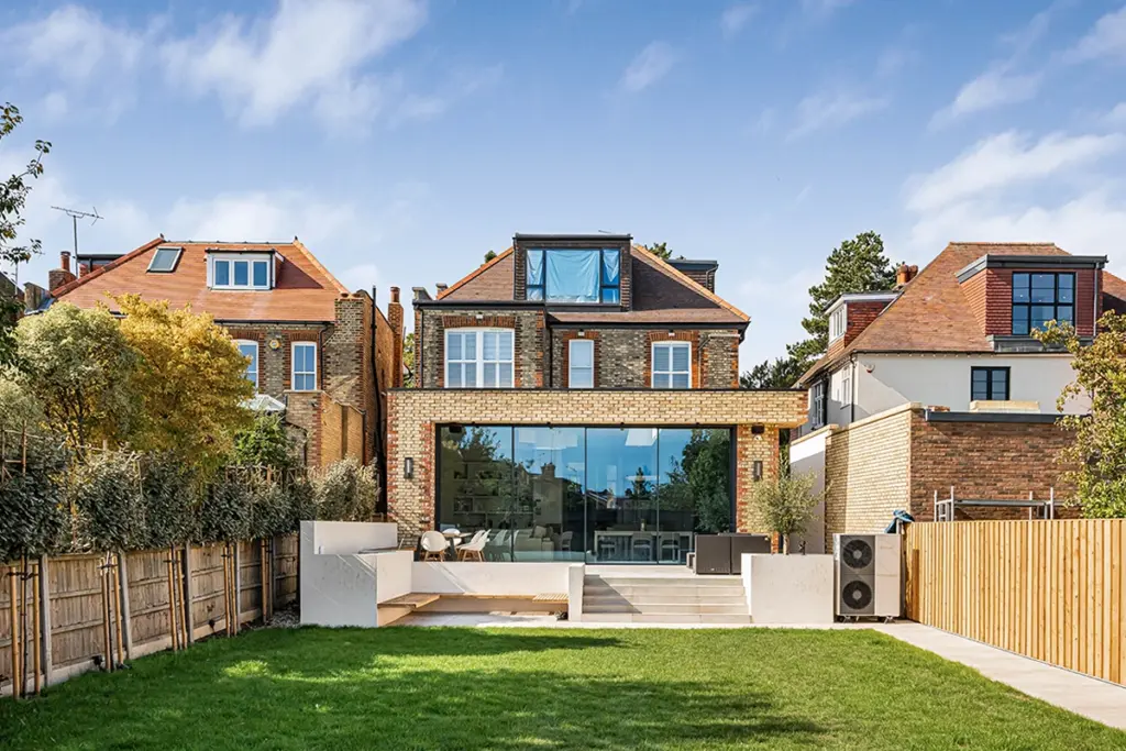 A back garden with a lawn and stone patio leading to a brick house featuring a floor-to-ceiling glass wall extension.