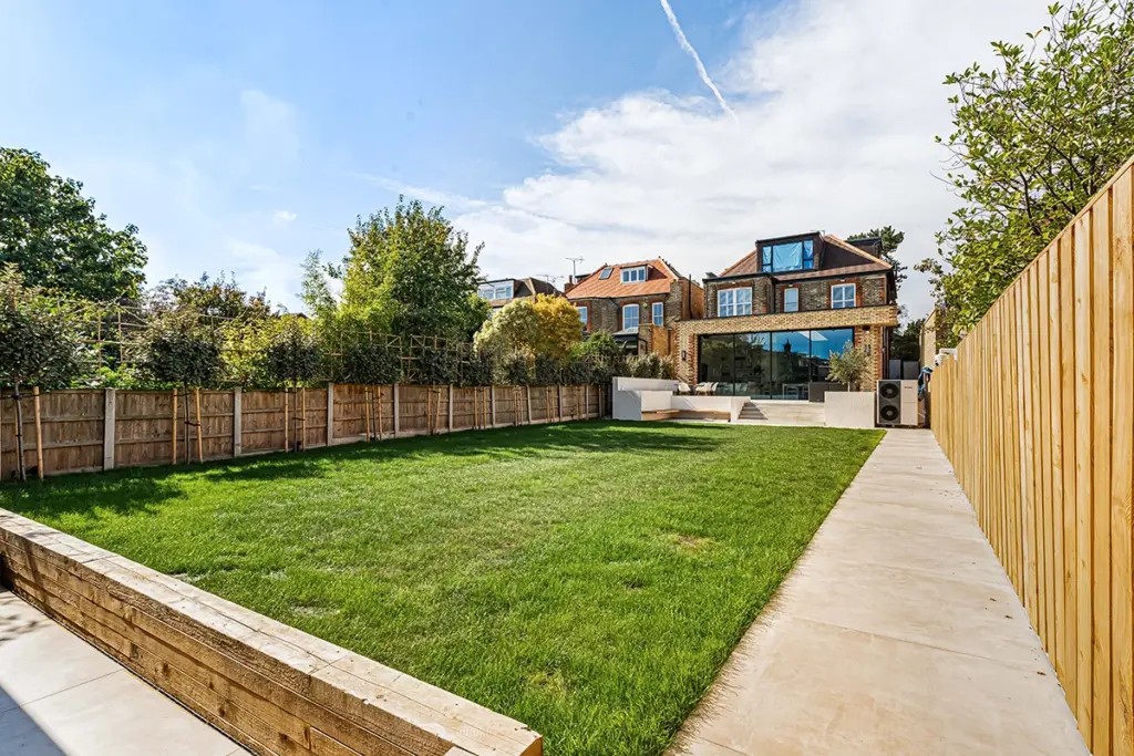 A large grassy back garden with a tiered timber retaining wall and a stone patio leading to a multi-storey house with a rear glass extension.