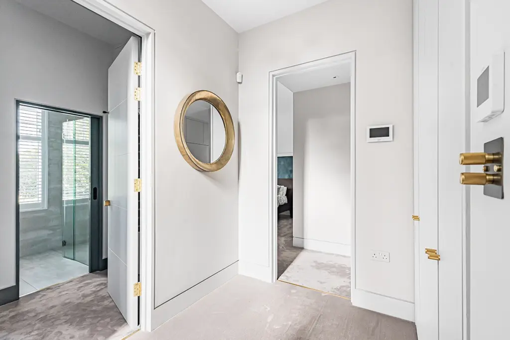 A hallway with beige carpet features a circular gold-rimmed mirror on the wall, an open doorway leading to a bathroom, and a second doorway leading to a bedroom.