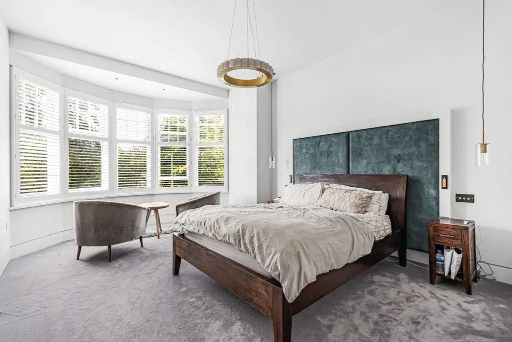 A bedroom featuring a dark wood bed frame with a large blue upholstered headboard, two grey armchairs by the bay window, and a modern circular pendant light.
