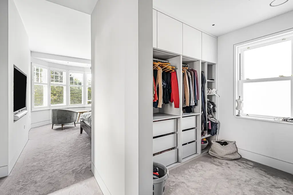 A walk-through wardrobe area with open shelving and hanging clothes, adjacent to a bedroom with a view of a bay window.