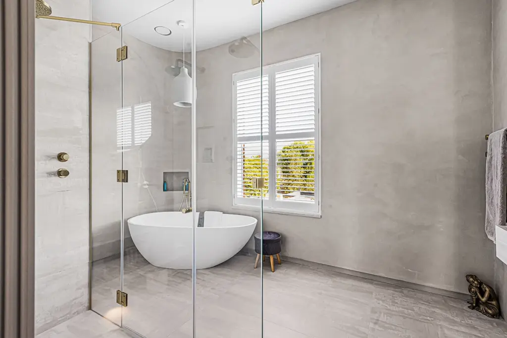 A glass-enclosed shower area featuring a white freestanding bath, a small stool, and a window with white shutters.