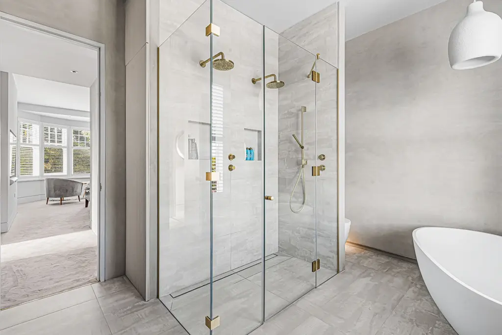 A glass-enclosed walk-in shower with gold fittings and a recessed shelf, next to a freestanding white bath in a modern bathroom.