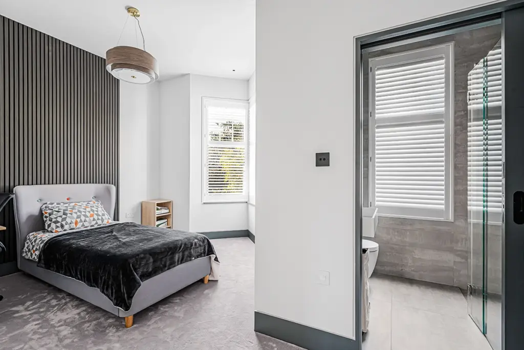 A modern bedroom with a grey bed against a slatted wall, leading into an en suite bathroom with a glass shower.