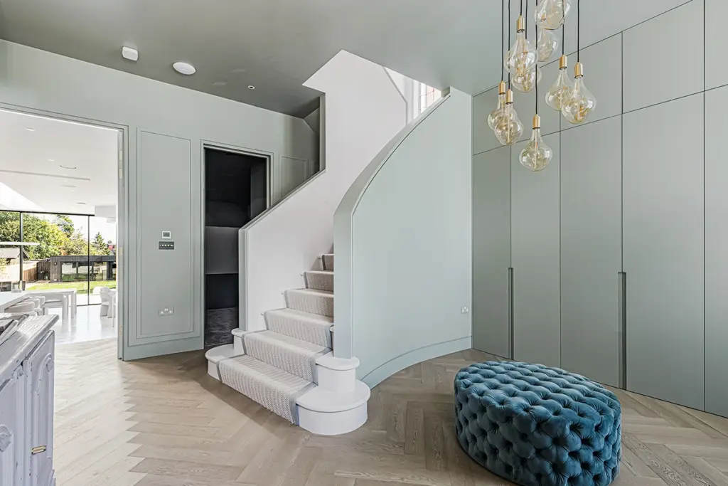 A modern entrance hall with a curved staircase, a blue tufted ottoman, and floor-to-ceiling sage green cupboards illuminated by a cluster of pendant lights.